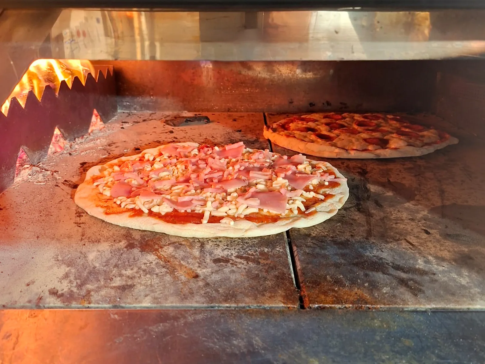Dos pizzas cociéndose en el horno de leña de Pizzería El 26, con la llama encendida a un lado